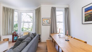 A modern living room with a sofa, a wooden dining table, and a bench. There are curtains on the windows, and a framed picture on the wall. - Leafy & Stylish Brixton Retreat (London)