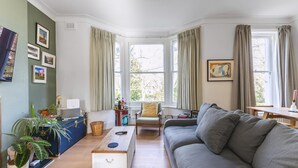 A cozy living room with a sofa, a small table, a chair, and a large window with curtains. - Leafy & Stylish Brixton Retreat (London)
