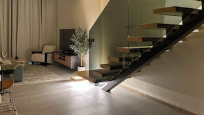 A modern living room with a glass staircase, a flat-screen TV, a potted plant, and a seating area. - Stylish 1BR Penthouse in The Autograph  (Dubai)
