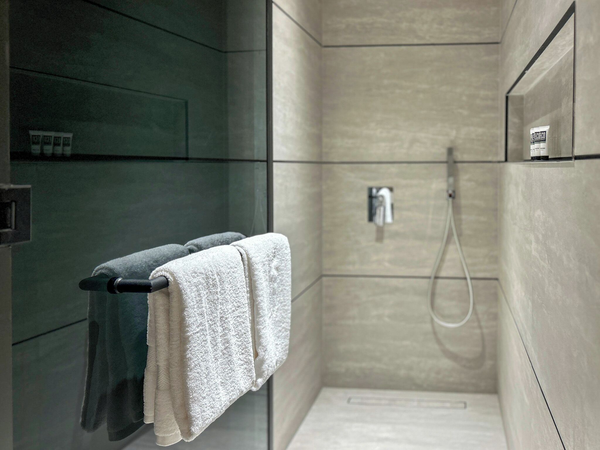 Een moderne douche met een glazen deur, een handdoekhouder met twee handdoeken en een kleine fles shampoo aan de muur.