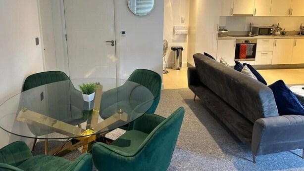 A modern living room with a glass dining table, green upholstered chairs, a grey sofa, and a kitchen area with white cabinets and a microwave. - DH Aparthotels - Cromwell Apartment (Huntingdon)