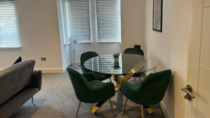 A modern living room with a glass-top coffee table, green upholstered chairs, a grey sofa, and a large window with blinds. - DH Aparthotels - Cromwell Apartment (Huntingdon)