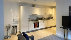 A modern kitchen with white cabinets, a microwave, stove, and a sink. There is a small dining area with a table and chairs. - DH Aparthotels - Cromwell Apartment (Huntingdon)