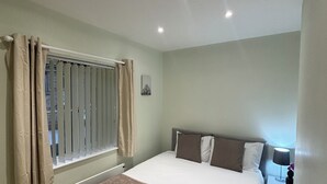 Un dormitorio con cama, mesita de noche, lámpara y ventana con cortinas.