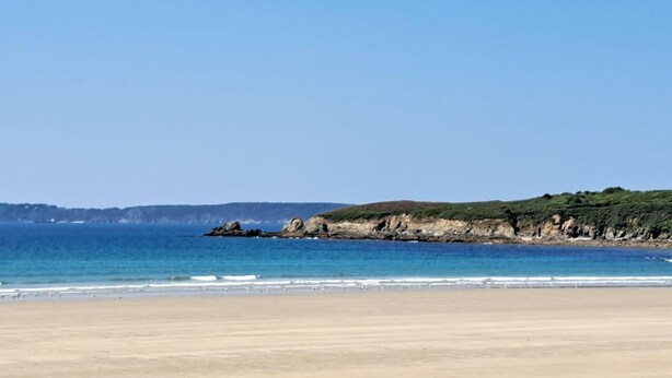 Ein Strand mit blauem Himmel, ruhigem Wasser und einer felsigen Küste.
