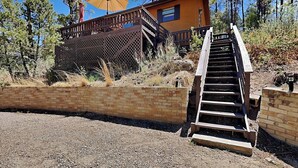 A wooden staircase leading to a cabin with a deck, surrounded by trees and a clear sky. - Knotty Pines Cabin | Pet-Friendly, Great Location (Ruidoso)