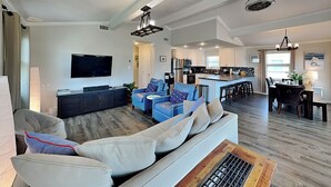 A modern living room with a large sofa, a coffee table, and a TV. There is a kitchen area with a dining table and chairs.