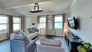A modern living room with a sofa, two armchairs, a coffee table, a TV, and a large window with curtains.
