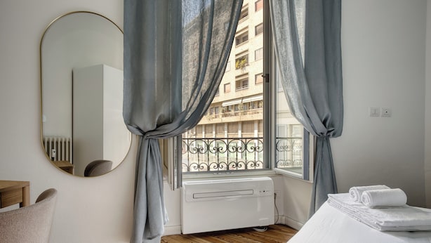 A hotel room with a wooden floor, a bed with white linens, a round mirror, and a window with curtains. - Classy apartment near Duomo di Milano (Milan)
