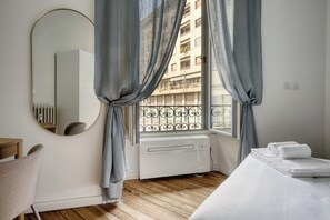 Une chambre d’hôtel avec un plancher en bois, un lit avec du linge de maison blanc, un miroir rond et une fenêtre avec des rideaux.