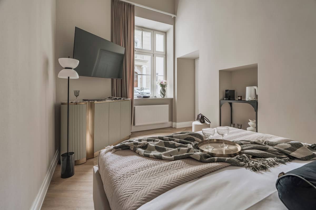 A modern hotel room with a large bed, a flat-screen TV, a bedside table with a lamp, a window with curtains, and a view of the outside.