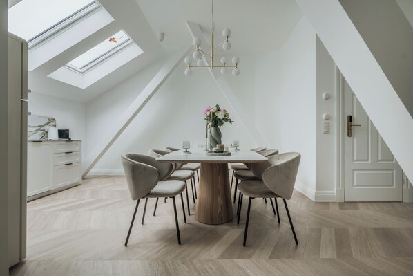 A modern dining area with a round table, six chairs, and a chandelier. A skylight and a door with a handle are visible. - La Belle Epoque (Warsaw)