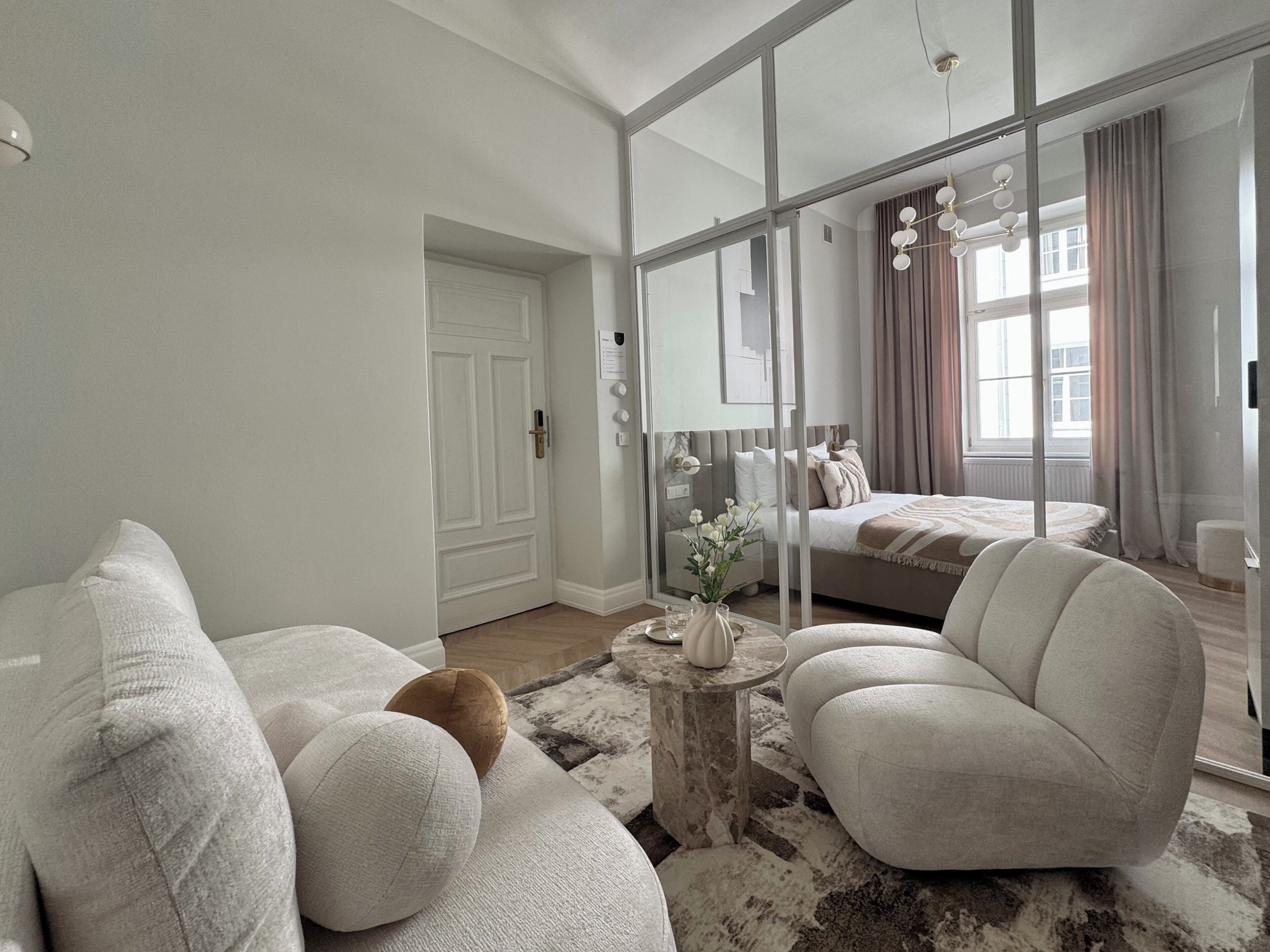 Ein modernes Wohnzimmer mit Sofa, Sesseln und Couchtisch. Durch eine Glastür ist ein Schlafzimmer mit Bett und Fenster sichtbar.