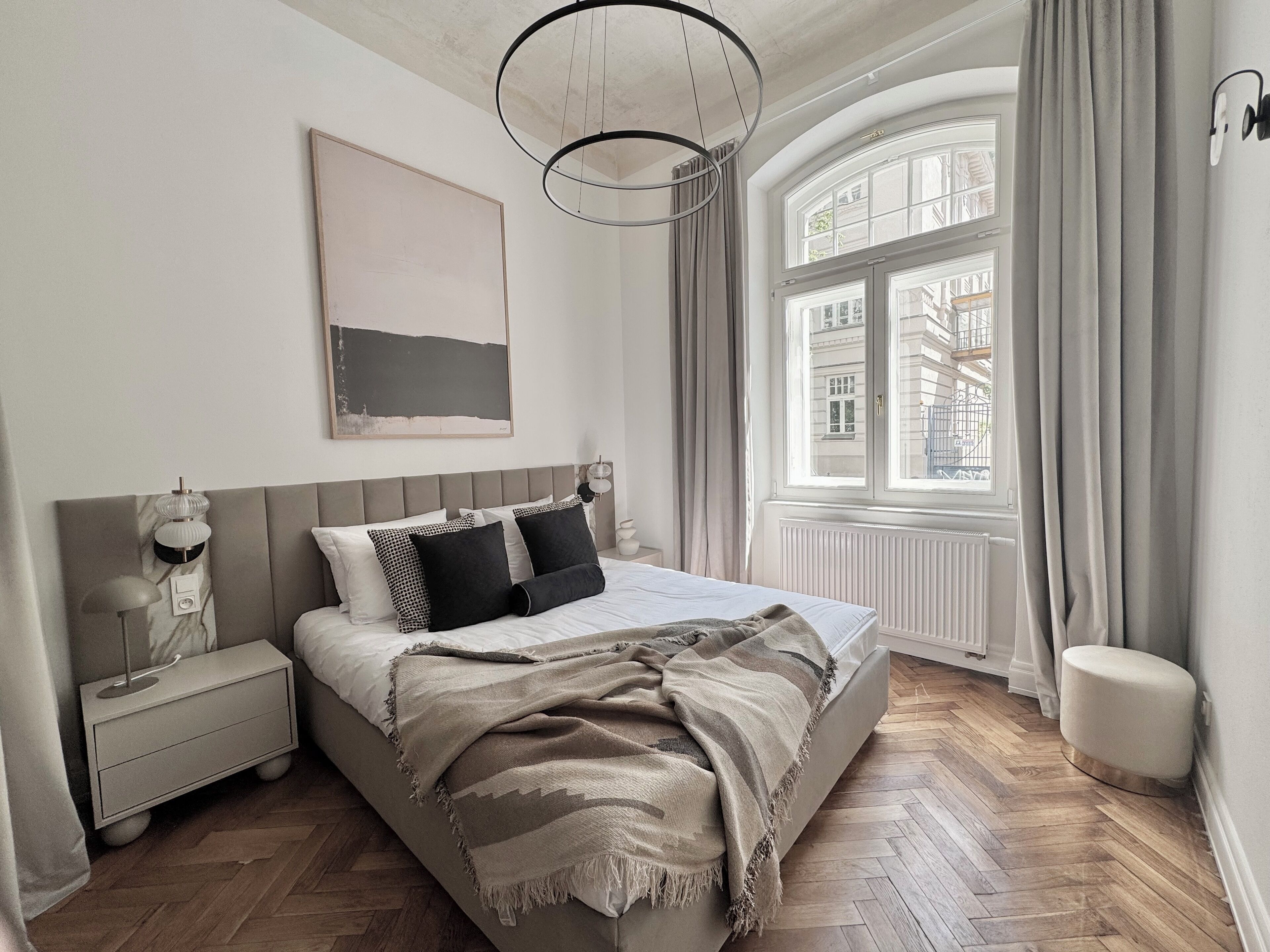 A modern bedroom with a large bed, a bedside table, a nightstand, a hanging light fixture, a window with curtains, and a radiator.