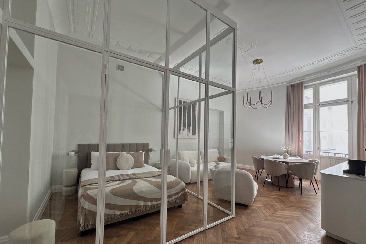A modern hotel room with a large bed, a glass partition, a dining area, and a view of the city.