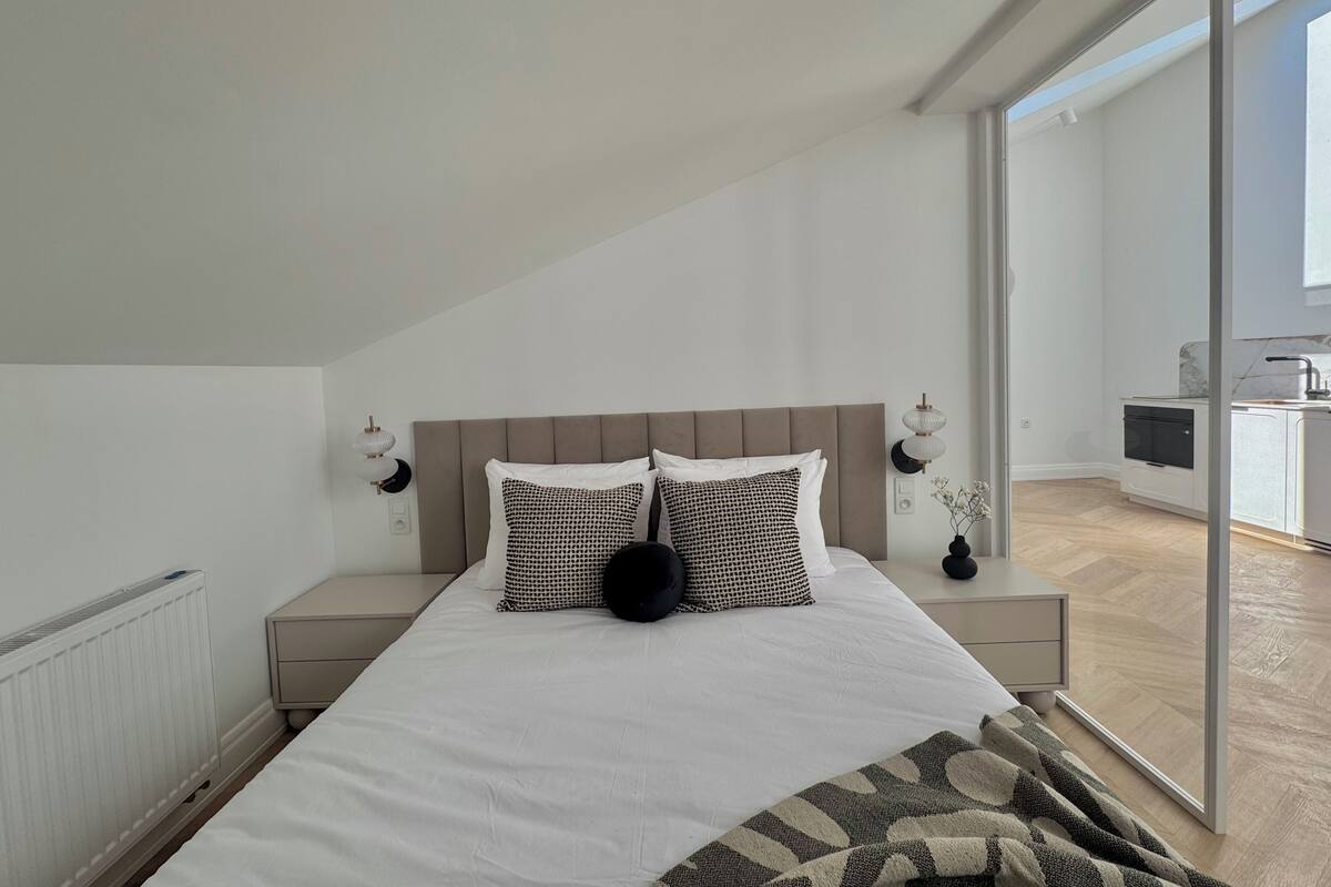A bedroom with a bed, bedside tables, a radiator, and a view of a kitchen and balcony.