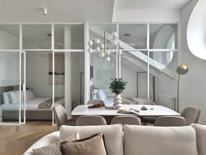 A modern living room with a large glass wall, a dining area with a white table, and a staircase leading to an upper level.