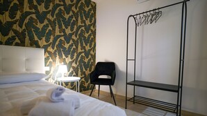 Ein ordentlich bezogenes Bett, ein aufgerolltes Handtuch, ein schwarzer Stuhl, ein kleiner Tisch mit einer Lampe und ein Metallschrank.