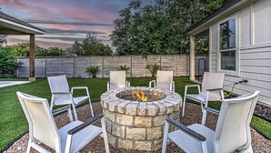 A stone fire pit with chairs around it, set in a backyard with a wooden fence and trees in the background.