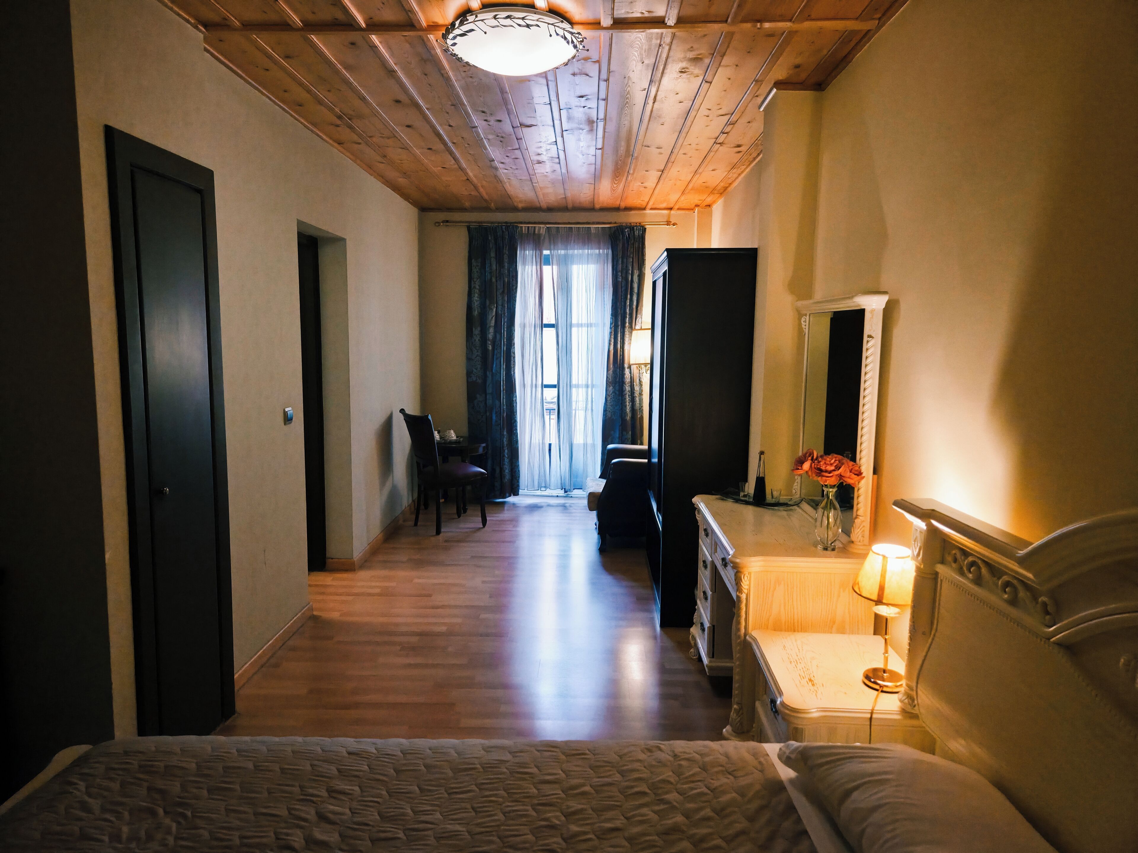 A hotel room with a wooden ceiling, a bed, a dresser, a mirror, and a window with curtains.
