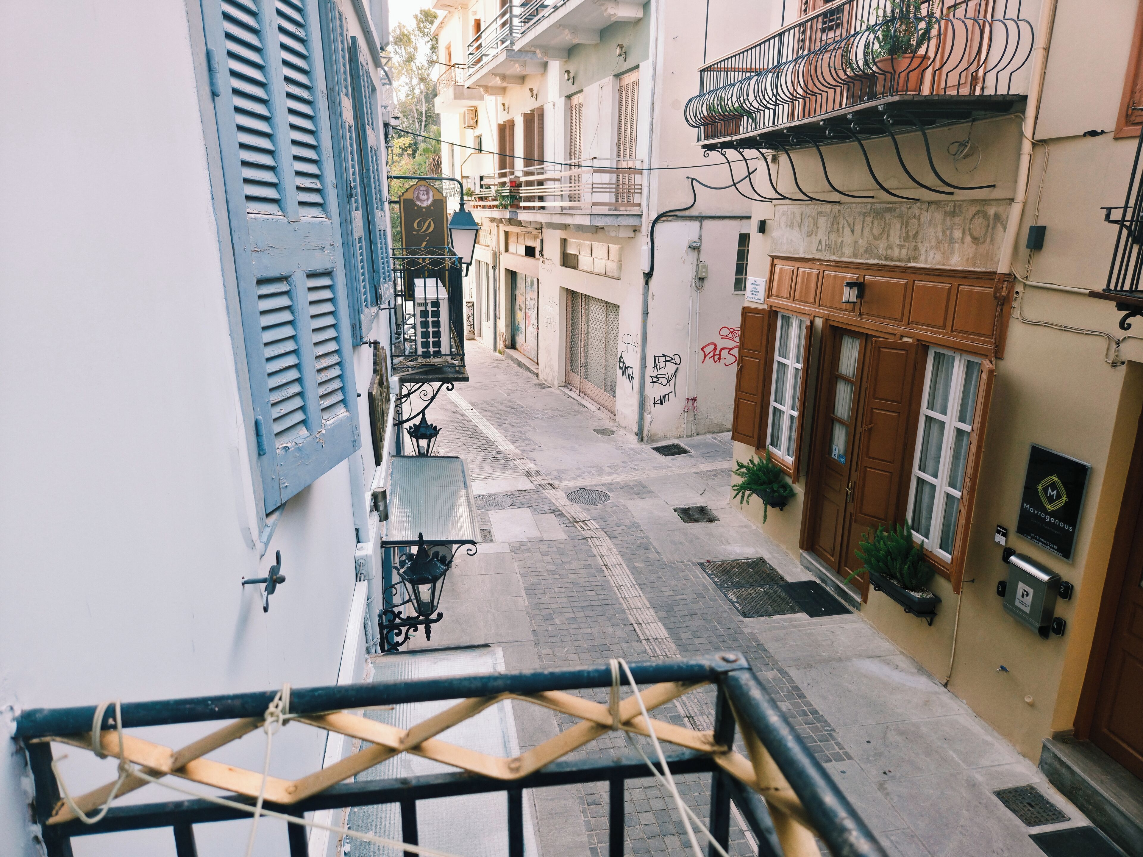 A narrow street with buildings on either side, featuring balconies with plants and outdoor seating.