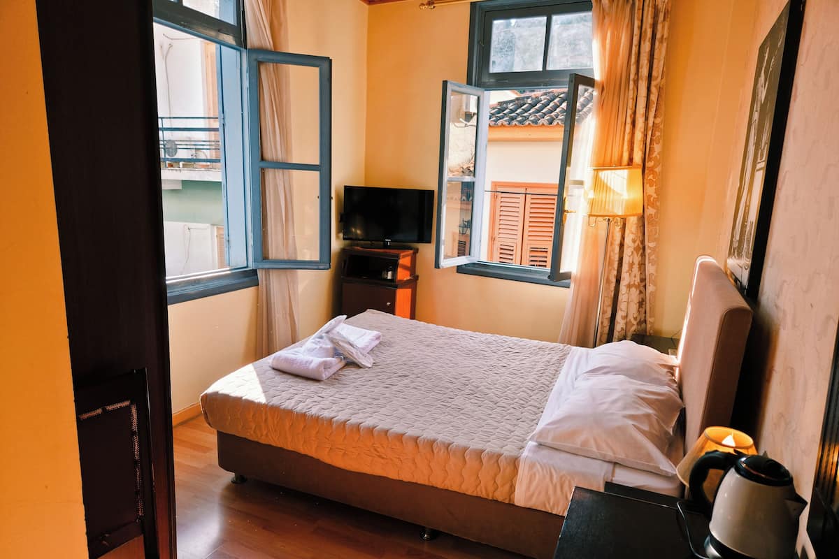 A neatly made bed with pillows and a folded towel, a bedside table with a kettle, a TV on a stand, and a window with curtains.