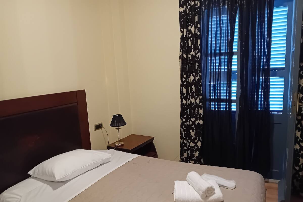A neatly made bed with white linens and a beige quilt, a bedside lamp, and a window with patterned curtains.