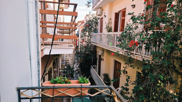 Une ruelle étroite en milieu urbain, bordée de bâtiments des deux côtés, avec des balcons ornés de plantes et de fleurs, et un portail en métal au premier plan.
