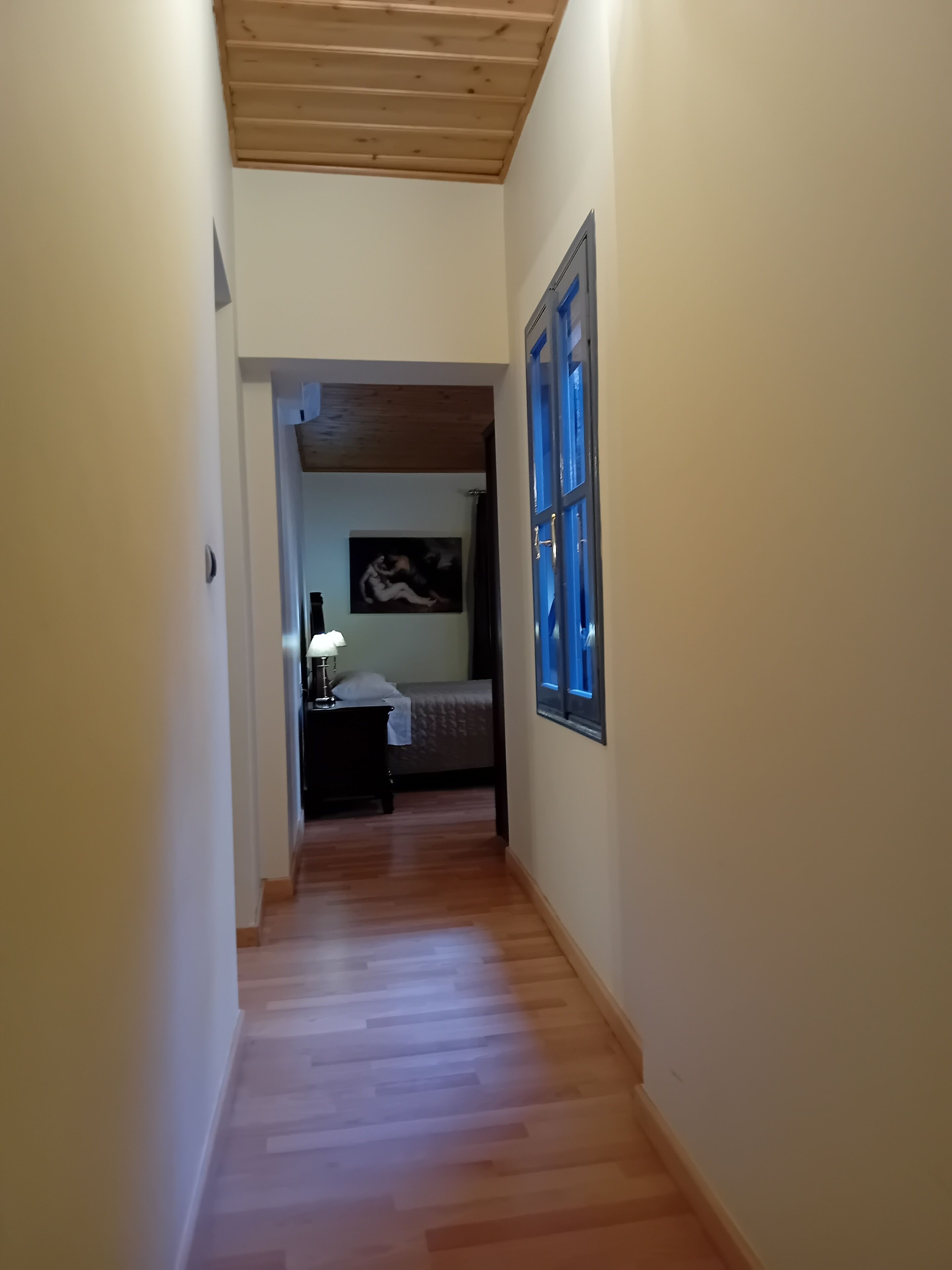 A narrow hallway with wooden flooring leading to a bedroom with a bed, a nightstand, and a painting on the wall.