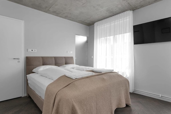 Un dormitorio con cama, una puerta, una ventana con cortinas y un televisor de pared.