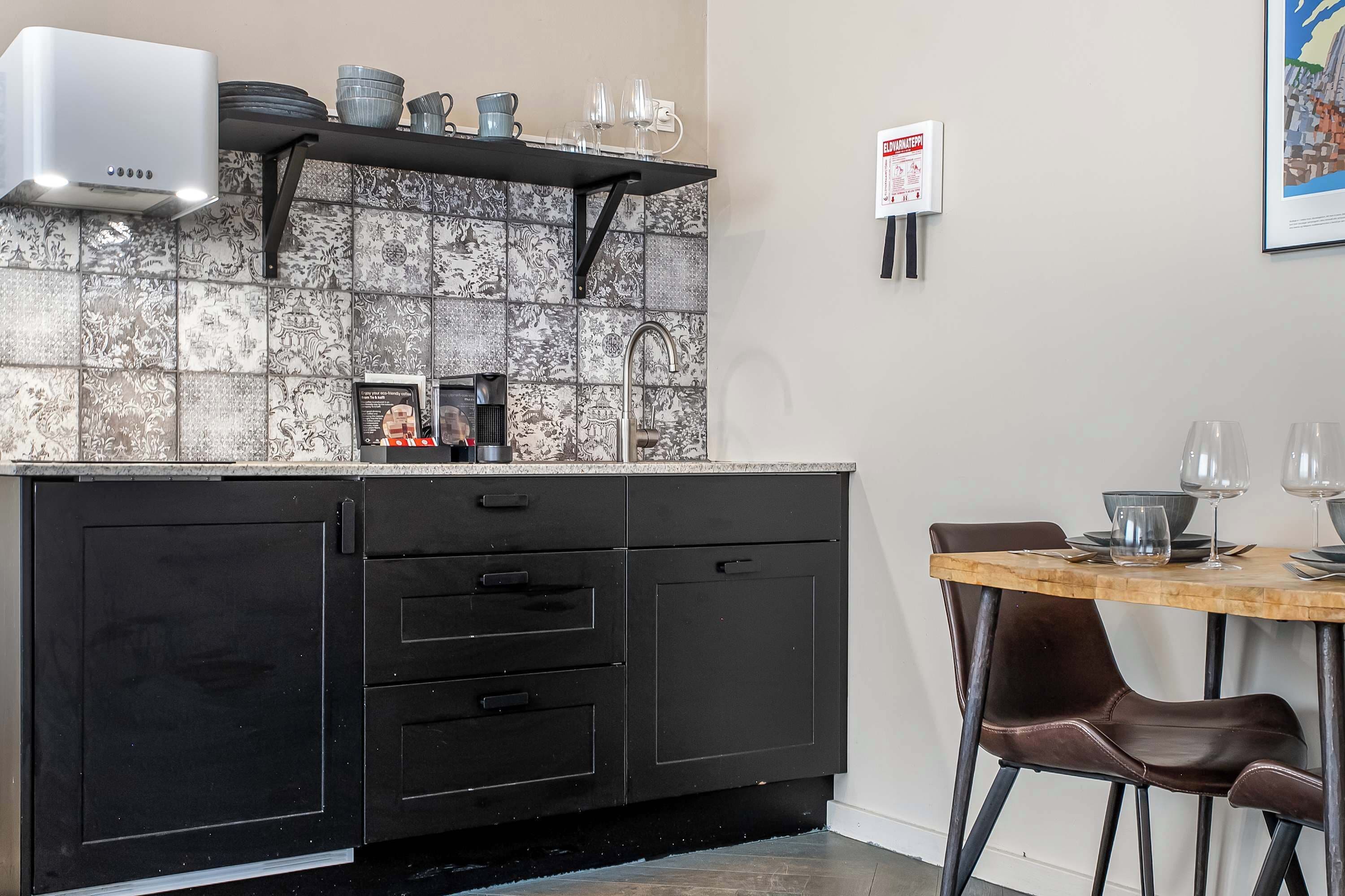 Cozinha compacta com bancada preta, cafeteira e painel de azulejos.