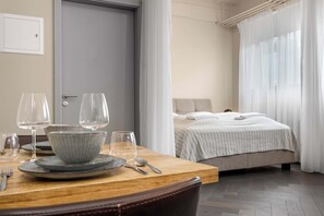 A neatly arranged dining area with a table set for two, a bed with a striped bedspread, and a window with sheer curtains.