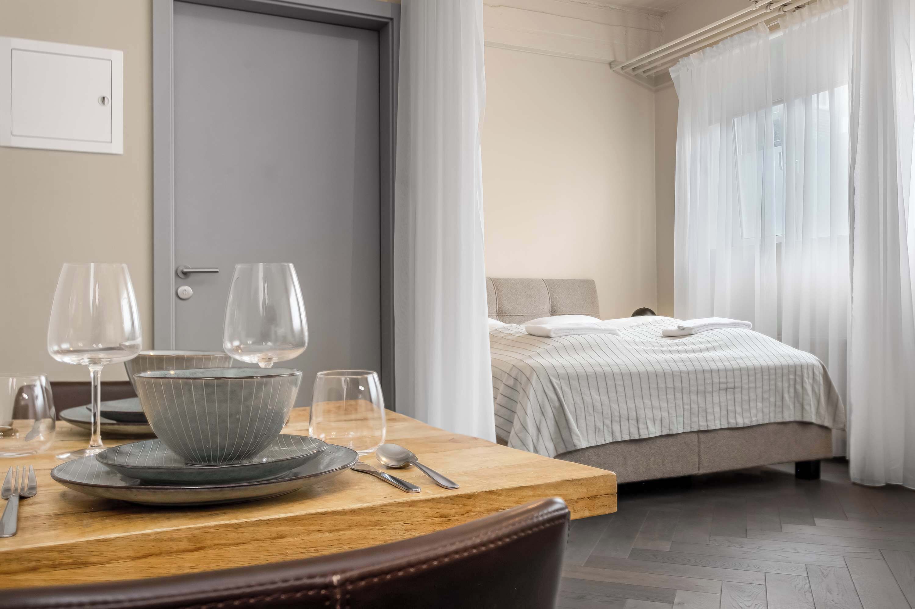 A neatly arranged dining area with a table set for two, a bed with a striped bedspread, and a window with sheer curtains.