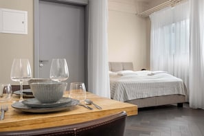 A neatly arranged dining area with a table set for two, a bed with a striped bedspread, and a window with sheer curtains.
