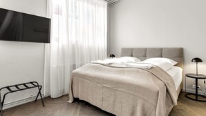 Un dormitorio moderno con una cama, un televisor instalado en la pared, una mesita de noche con una lámpara y un ventanal con cortinas translúcidas.