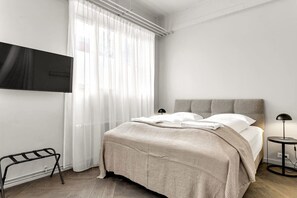 Une chambre moderne avec un lit, un téléviseur fixé au mur, une table de chevet avec une lampe et une fenêtre avec des rideaux légers.