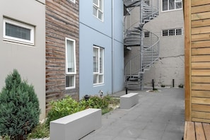 A narrow courtyard with a spiral staircase, a wooden fence, and a small garden area with plants and a bench.