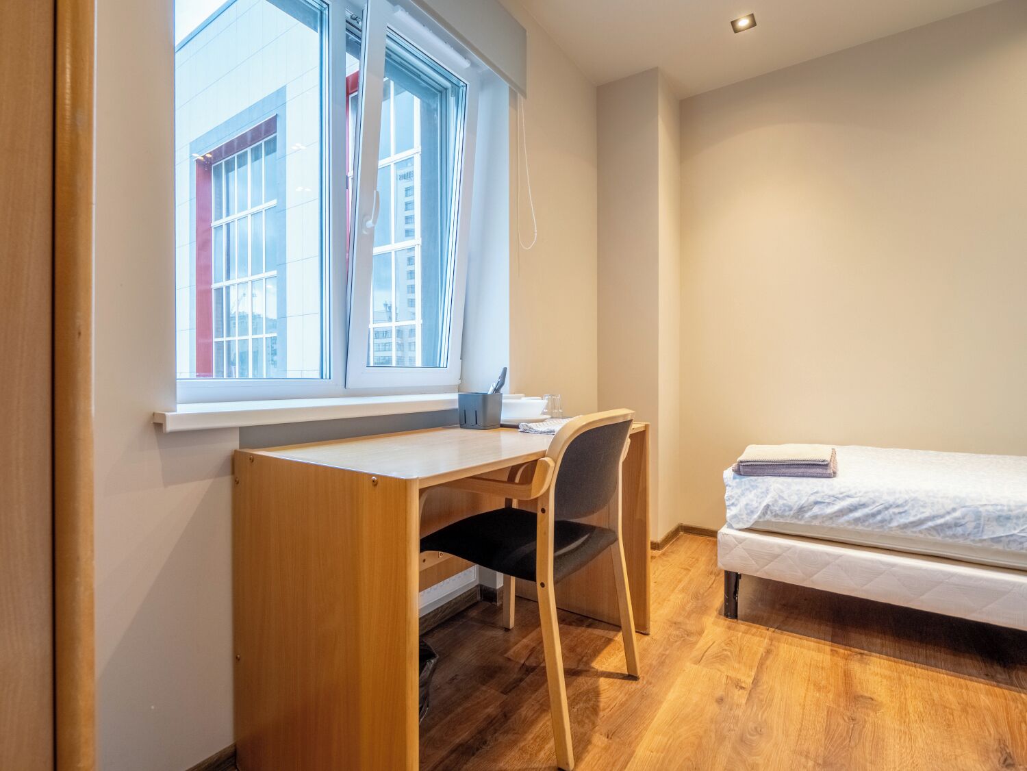 A small, well-lit room with a wooden desk and chair, a single bed, and a window with a view of buildings.