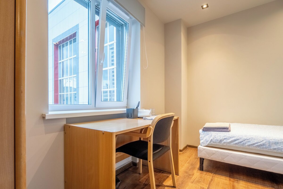 Una habitación pequeña y bien iluminada con un escritorio y una silla de madera, una cama individual y una ventana que ofrece vistas a edificios.