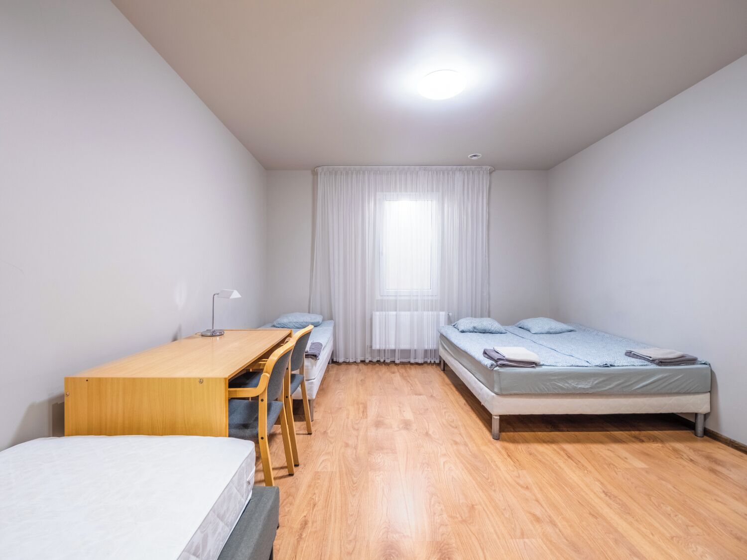 Habitació petita i ben il·luminada amb dos llits individuals, un escriptori de fusta amb una cadira i una finestra gran amb cortines translúcides.