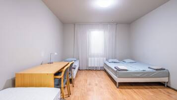 Habitació petita i ben il·luminada amb dos llits individuals, un escriptori de fusta amb una cadira i una finestra gran amb cortines translúcides.