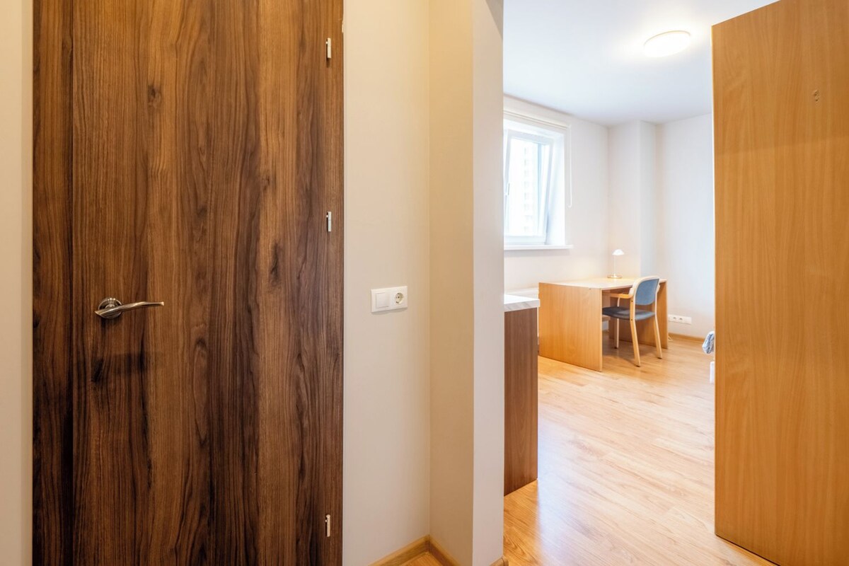 A modern apartment with a wooden door, a desk with a chair, and a window.