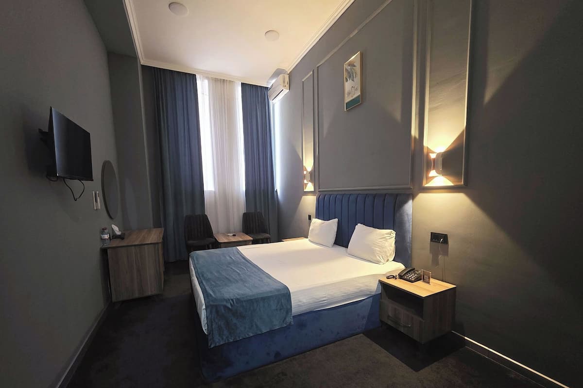 A hotel room with a large bed, a desk, a chair, a TV, and a bedside table with a lamp.