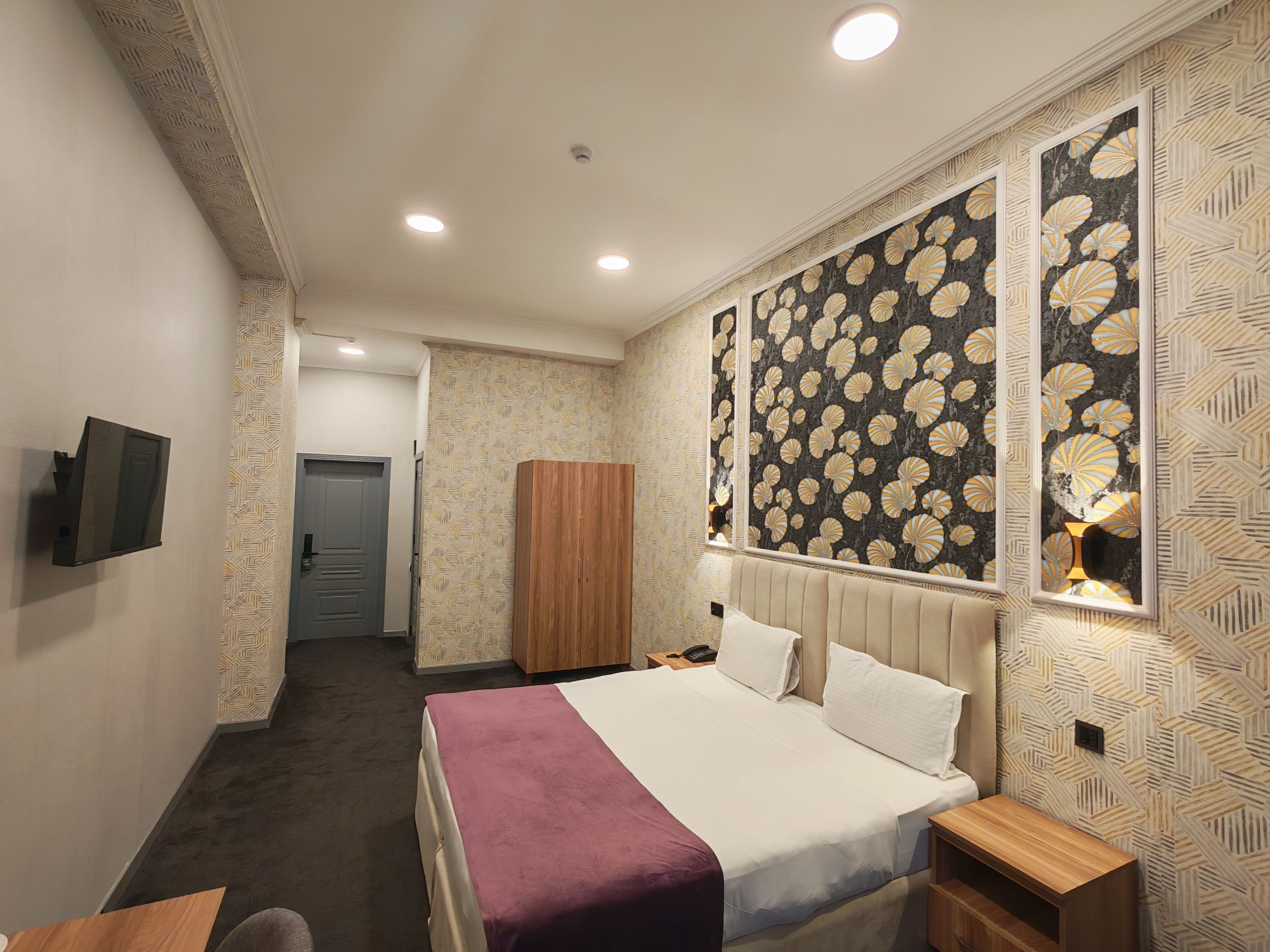 A hotel room with a bed, bedside tables, a wardrobe, a TV, and a patterned wall.