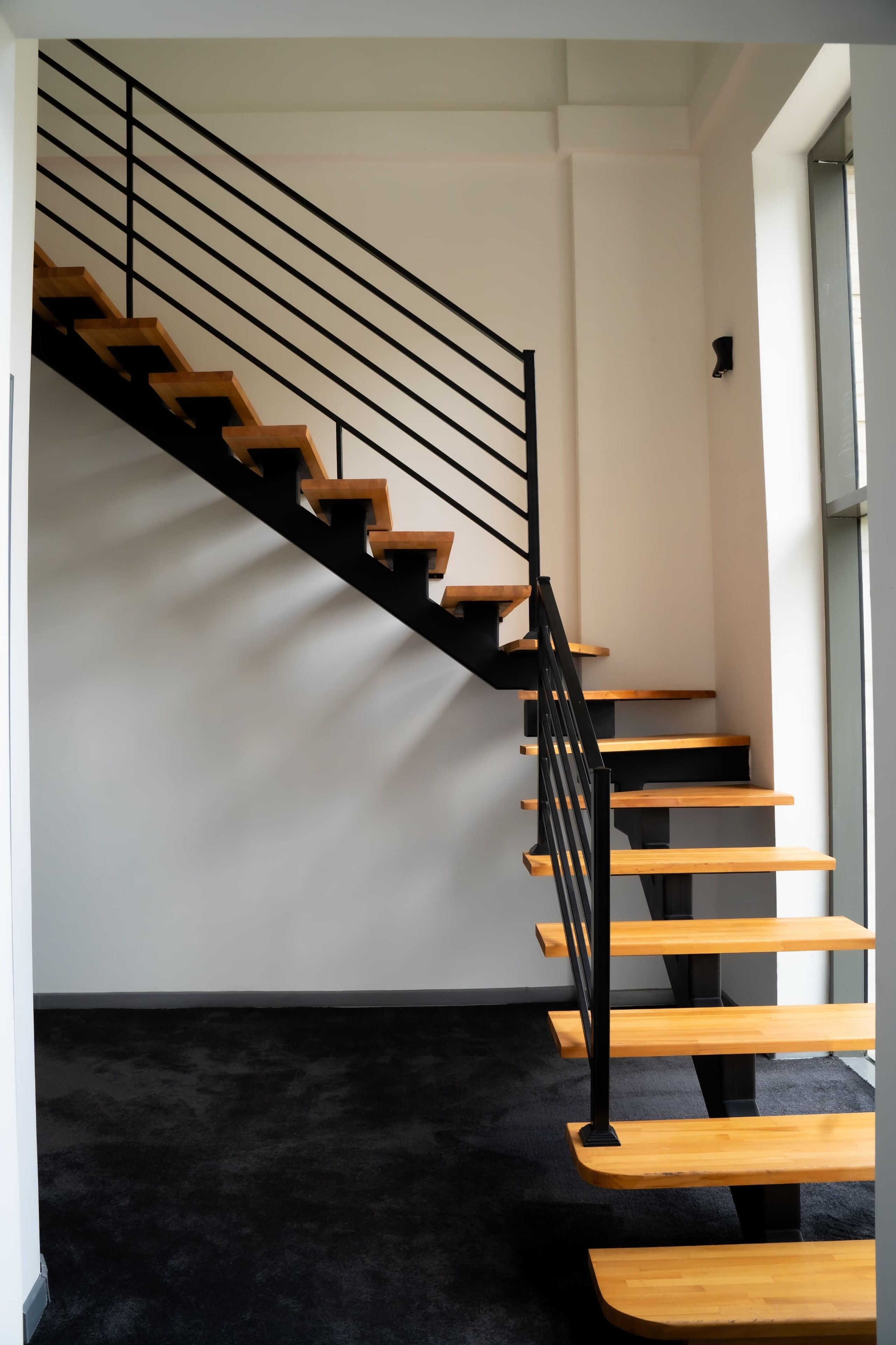A modern staircase with wooden steps and black metal railings, leading to a landing with a black carpeted floor.
