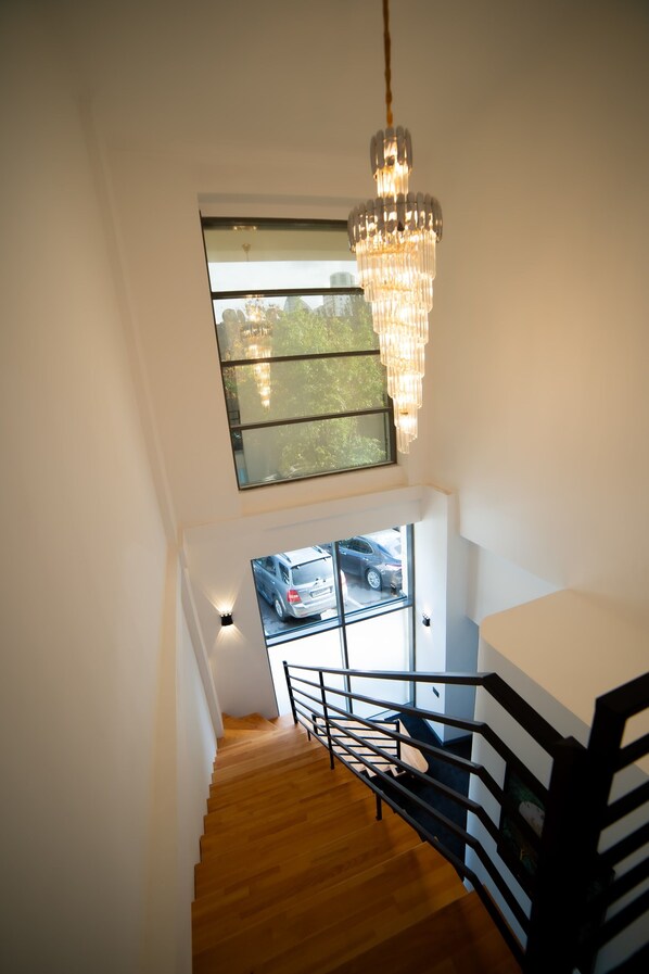 A staircase with wooden steps and black railings, leading to a room with a large window and a chandelier. - Will Hotel (Baku)
