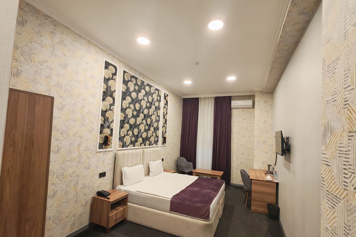 A hotel room with a bed, bedside tables, a desk, a chair, and an air conditioning unit.