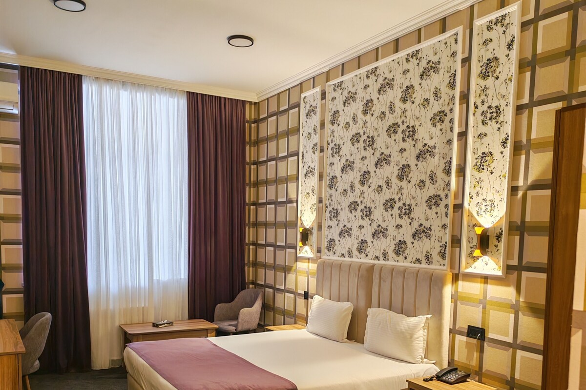 A hotel room with a large bed, a desk, a chair, a nightstand, and a window with curtains.