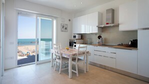 A modern kitchen with white cabinets, a dining table with chairs, a view of the beach through a glass door, and a small island with a microwave and coffee maker.