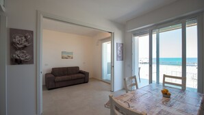 A spacious living room with a sofa, a dining table with chairs, and a view of the beach through large sliding glass doors.